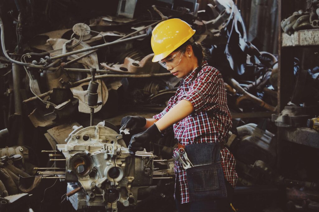 A female mechanic wearing a yellow hard hat and safety glasses is working on disassembling an engine in a cluttered workshop.