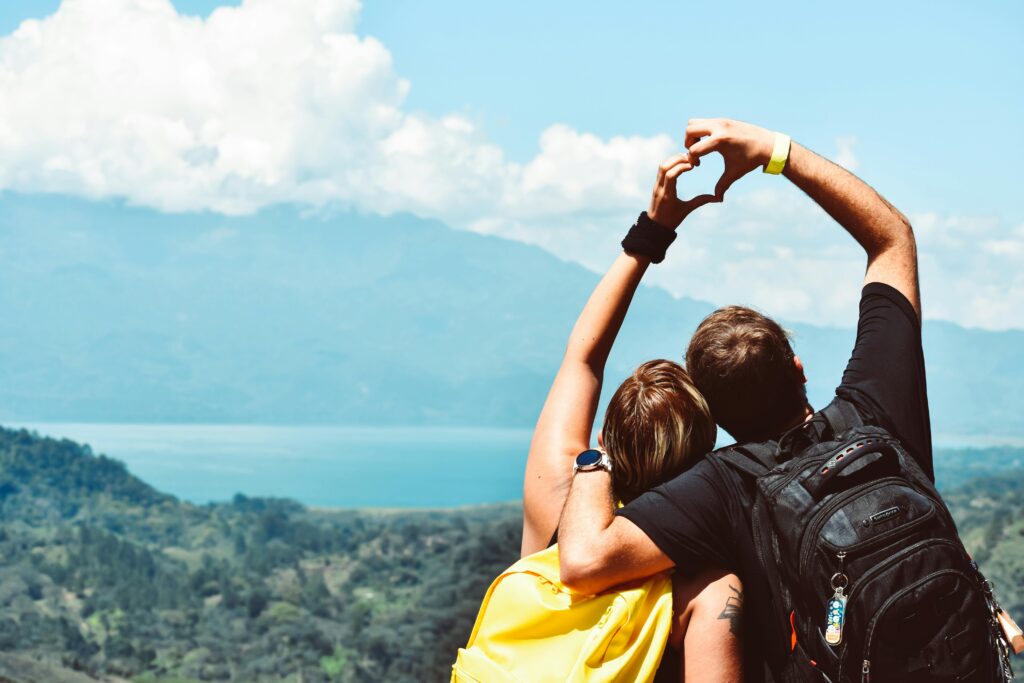A couple from behind, overlooking a scenic mountain landscape, forming a heart shape with their arms against a clear blue sky.