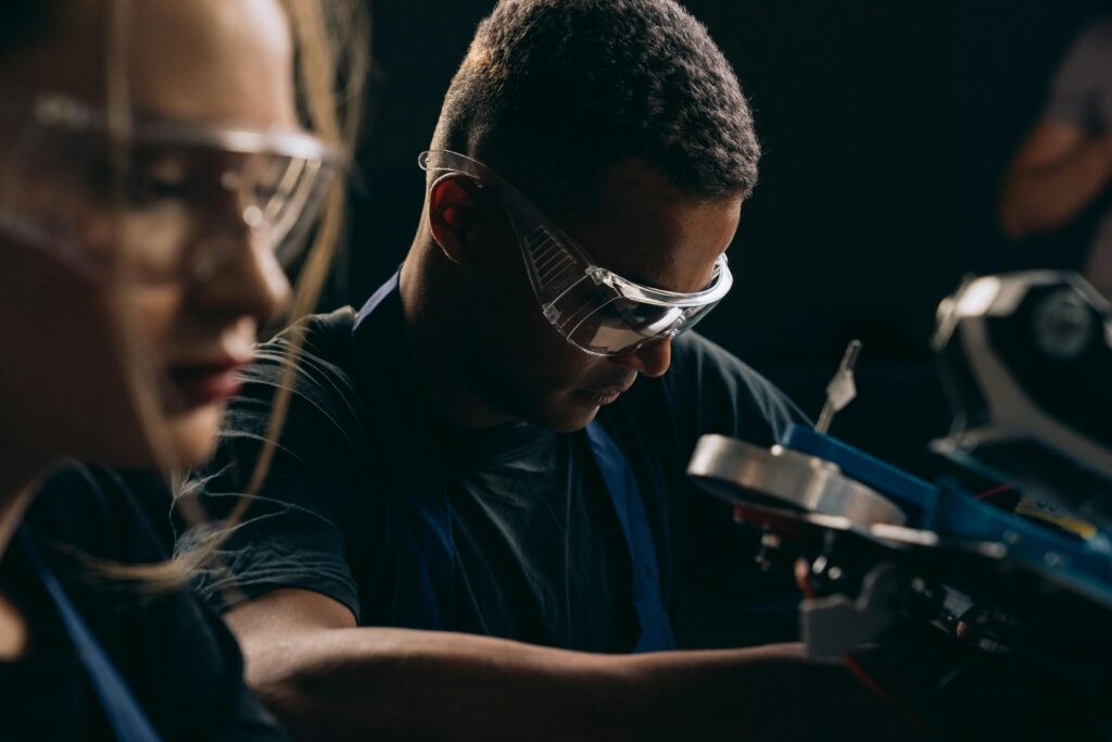 Two individuals wearing safety goggles and focused on a technical task. The person in the foreground is slightly out of focus, while the person in the background is working on machinery.