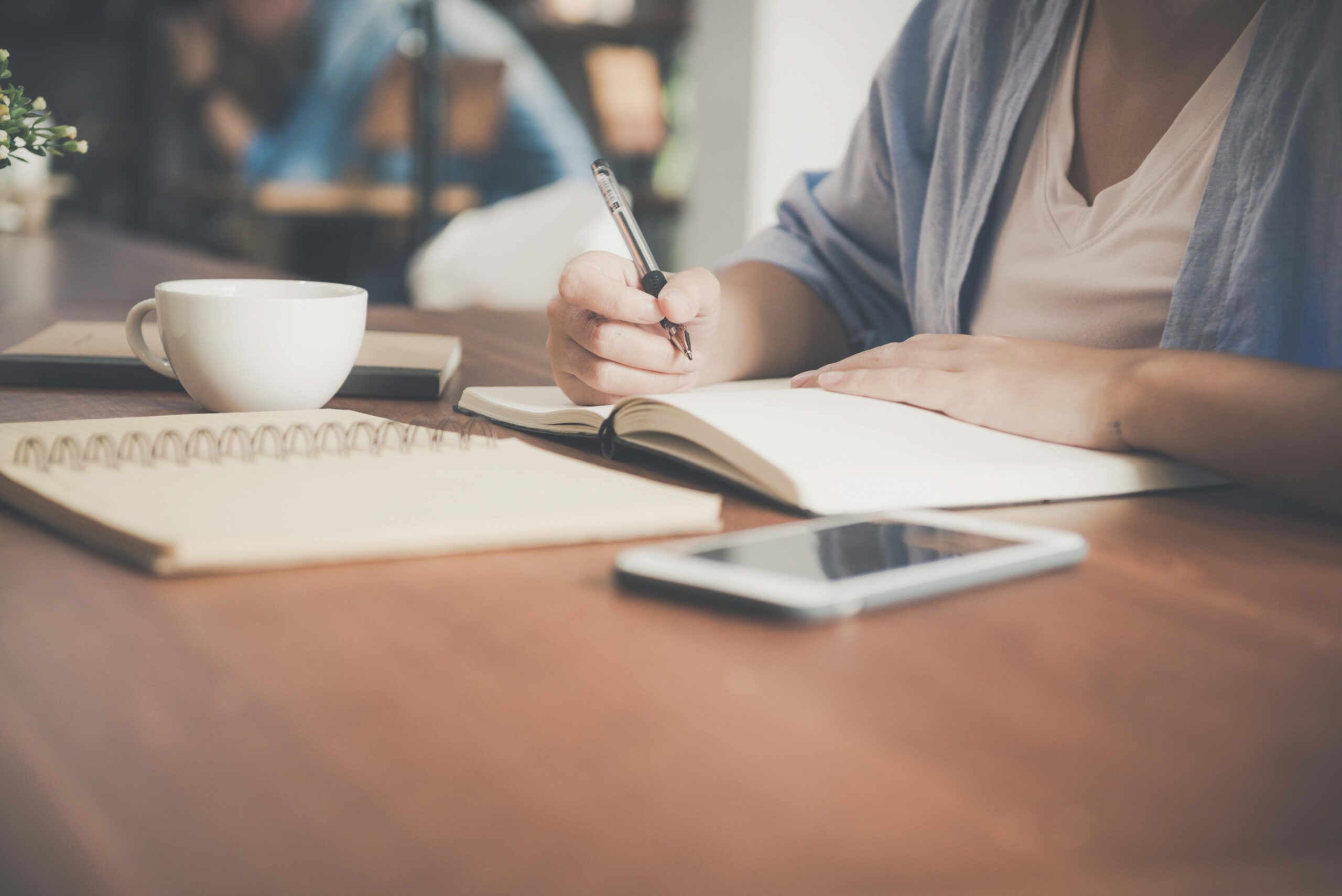 A person sitting at a wooden table in a cafe, writing in a notebook with a pen. There is a cup of coffee and a smartphone on the table, suggesting a setting conducive to work or study.