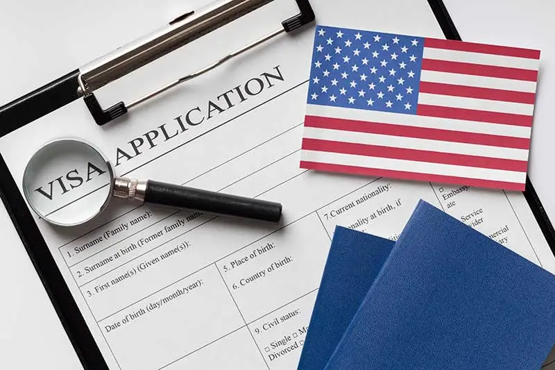Close-up of a U.S. visa application form on a clipboard, with a magnifying glass, two blue passports, and a small American flag, symbolizing the process of applying for a U.S. visa.