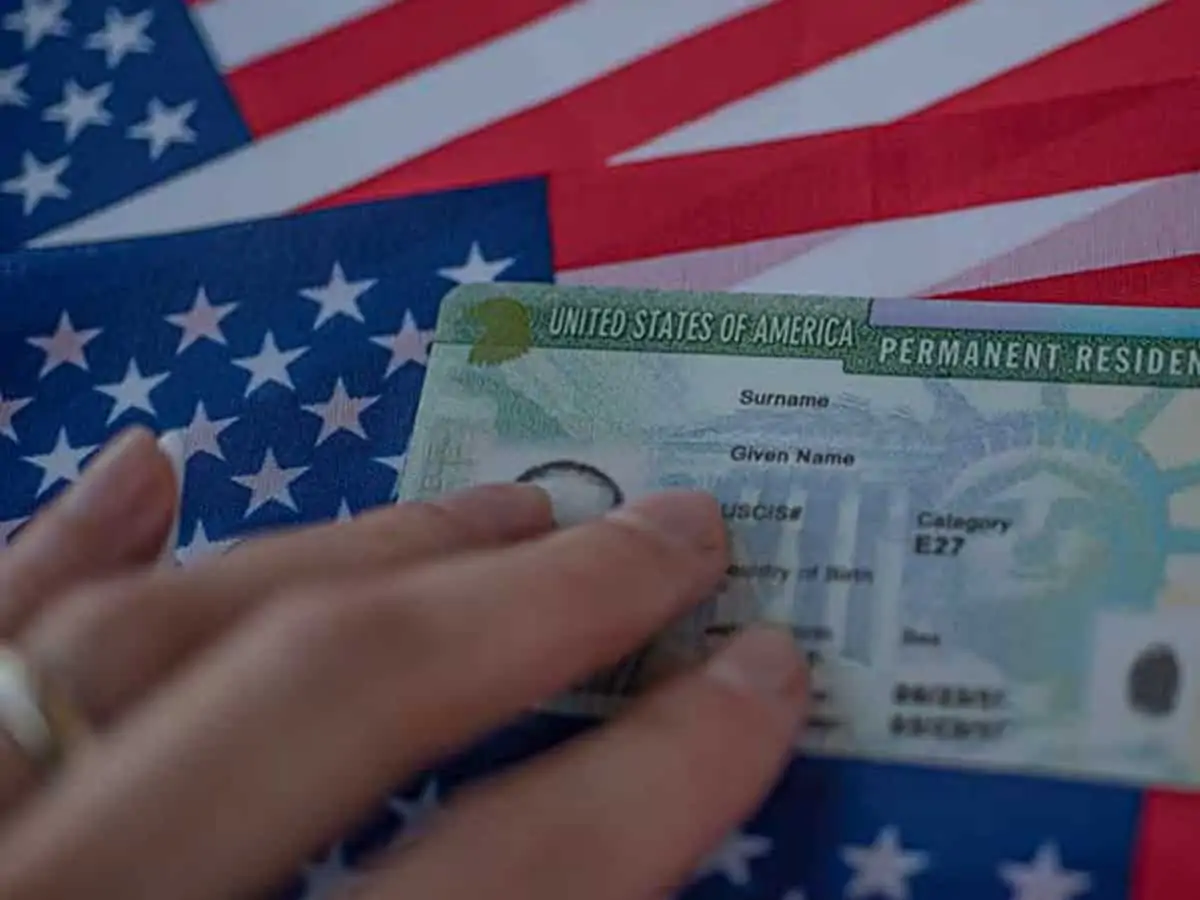 A hand holds a United States Permanent Resident Card (Green Card) over an American flag.