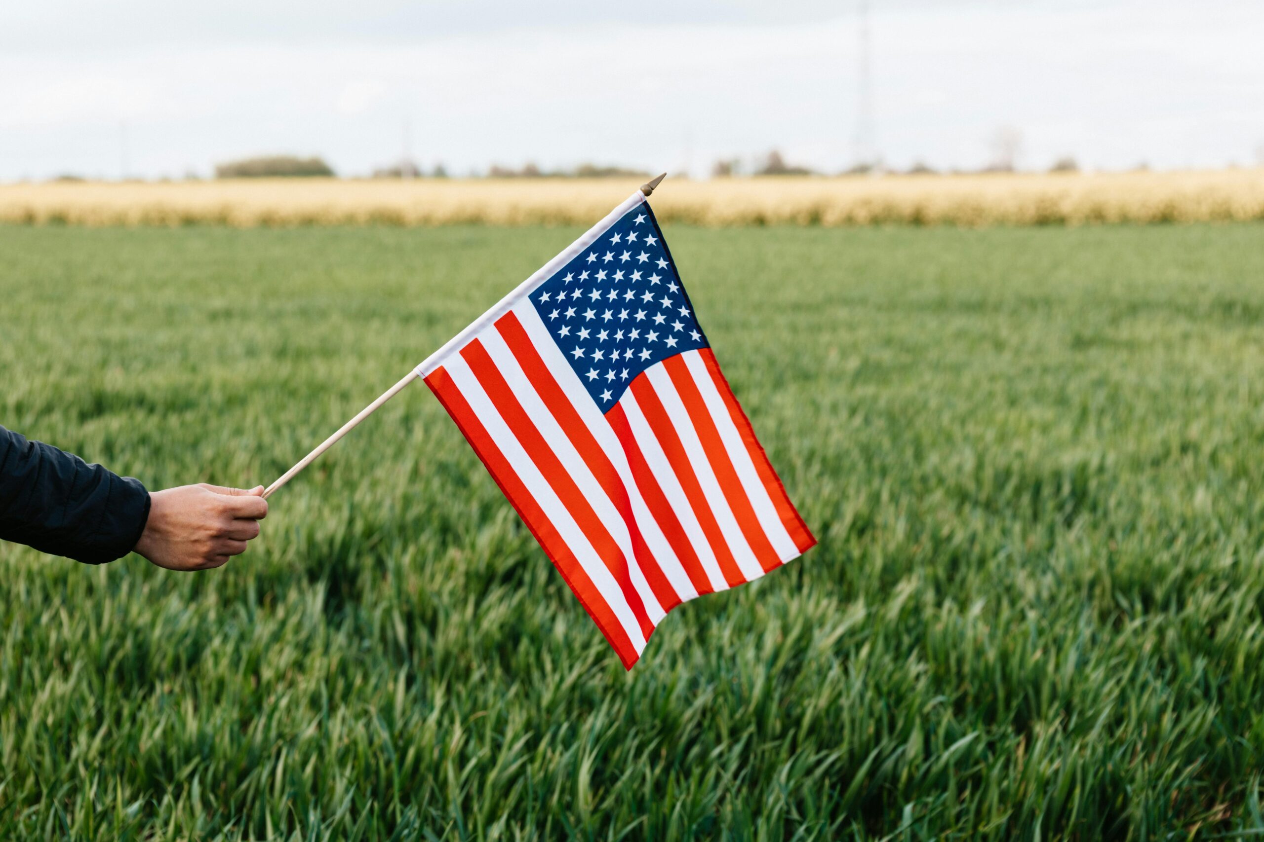 La imagen muestra una bandera estadounidense sostenida por una mano, ondeando contra un fondo de campo verde. La bandera es clara y vibrante, con sus franjas rojas y blancas y un campo de estrellas en un fondo azul. El entorno rural y la composición de la imagen capturan un sentido de patriotismo y tranquilidad natural.