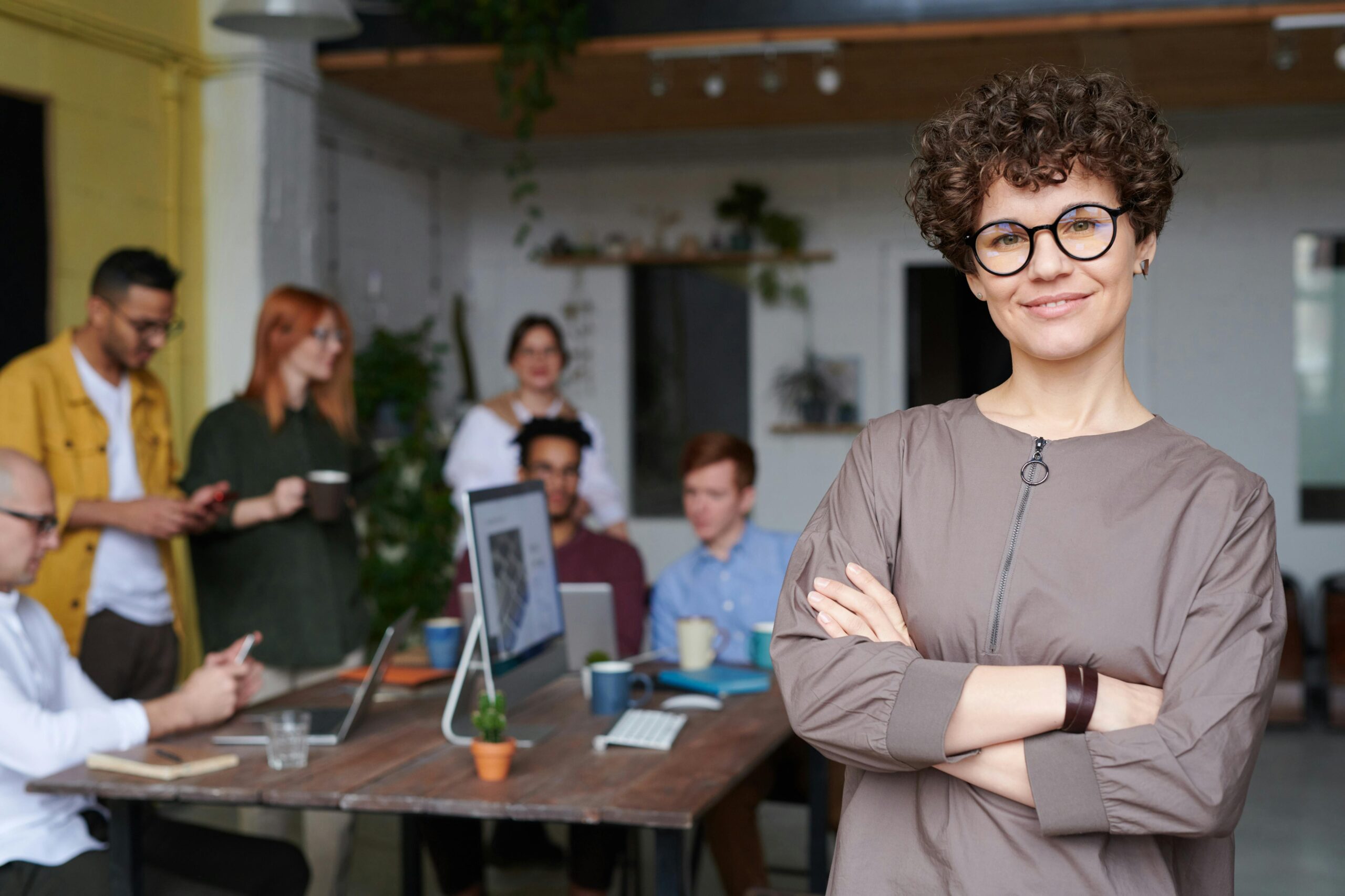 A confident person with curly hair and glasses is standing with arms crossed and smiling at the camera. In the background, a group of people is engaged in a casual meeting around a table with laptops and coffee cups in an open, modern office space.A confident person with curly hair and glasses is standing with arms crossed and smiling at the camera. In the background, a group of people is engaged in a casual meeting around a table with laptops and coffee cups in an open, modern office space.