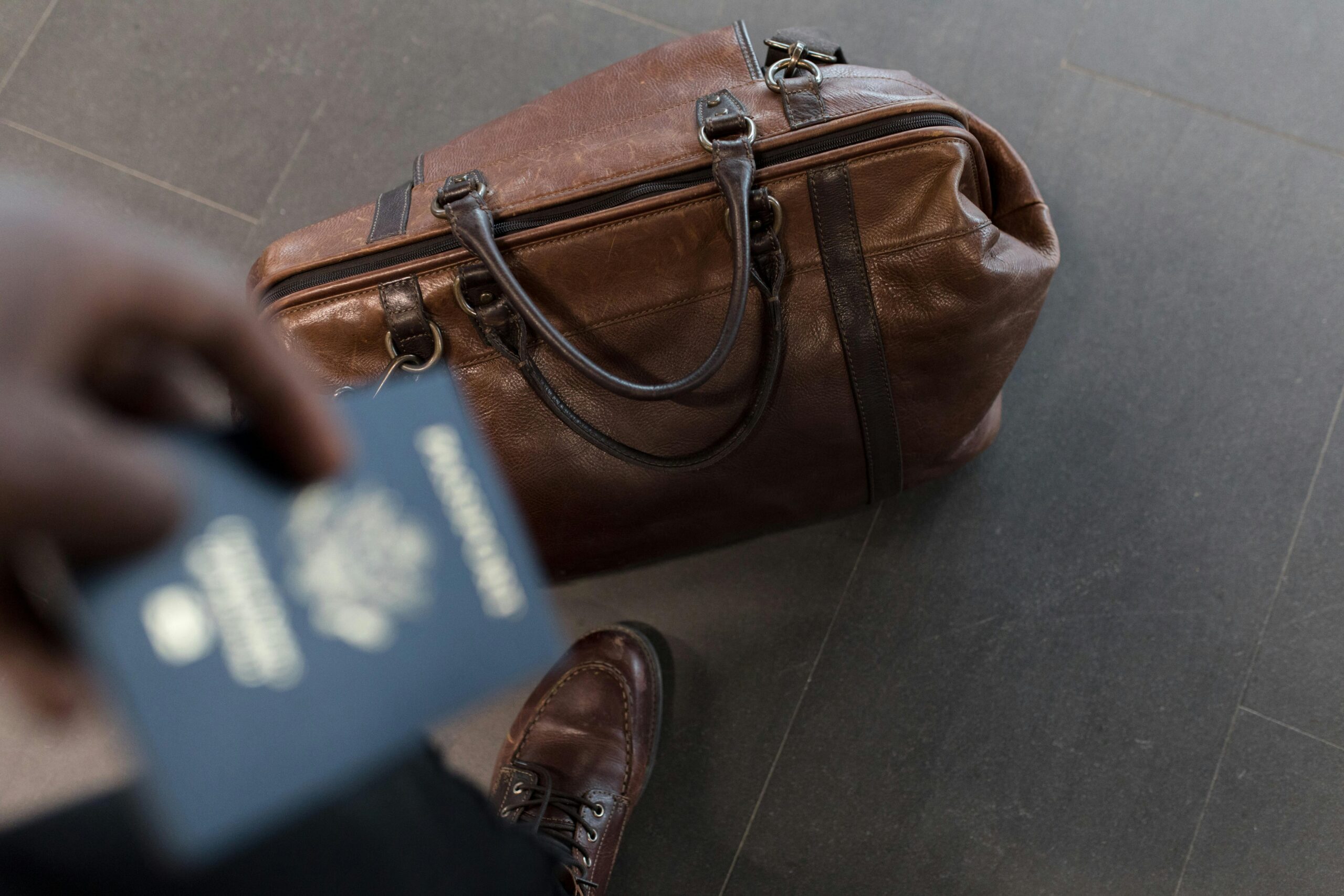 Primer plano de una persona sosteniendo un pasaporte, con una bolsa de viaje de cuero marrón y un zapato marrón en el fondo.