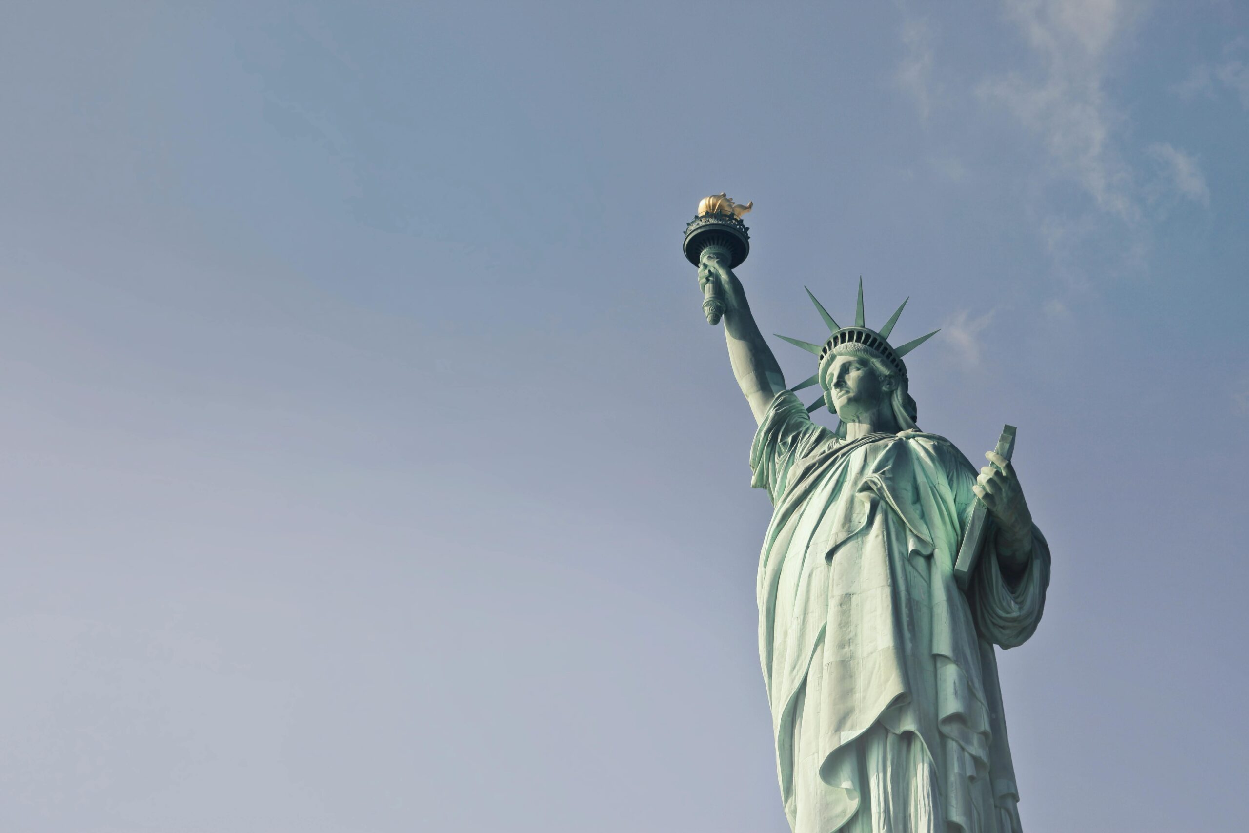 Statue of Liberty in New York, viewed from below with a clear blue sky in the background. The statue holds a torch high in its right hand and a tablet in its left hand.