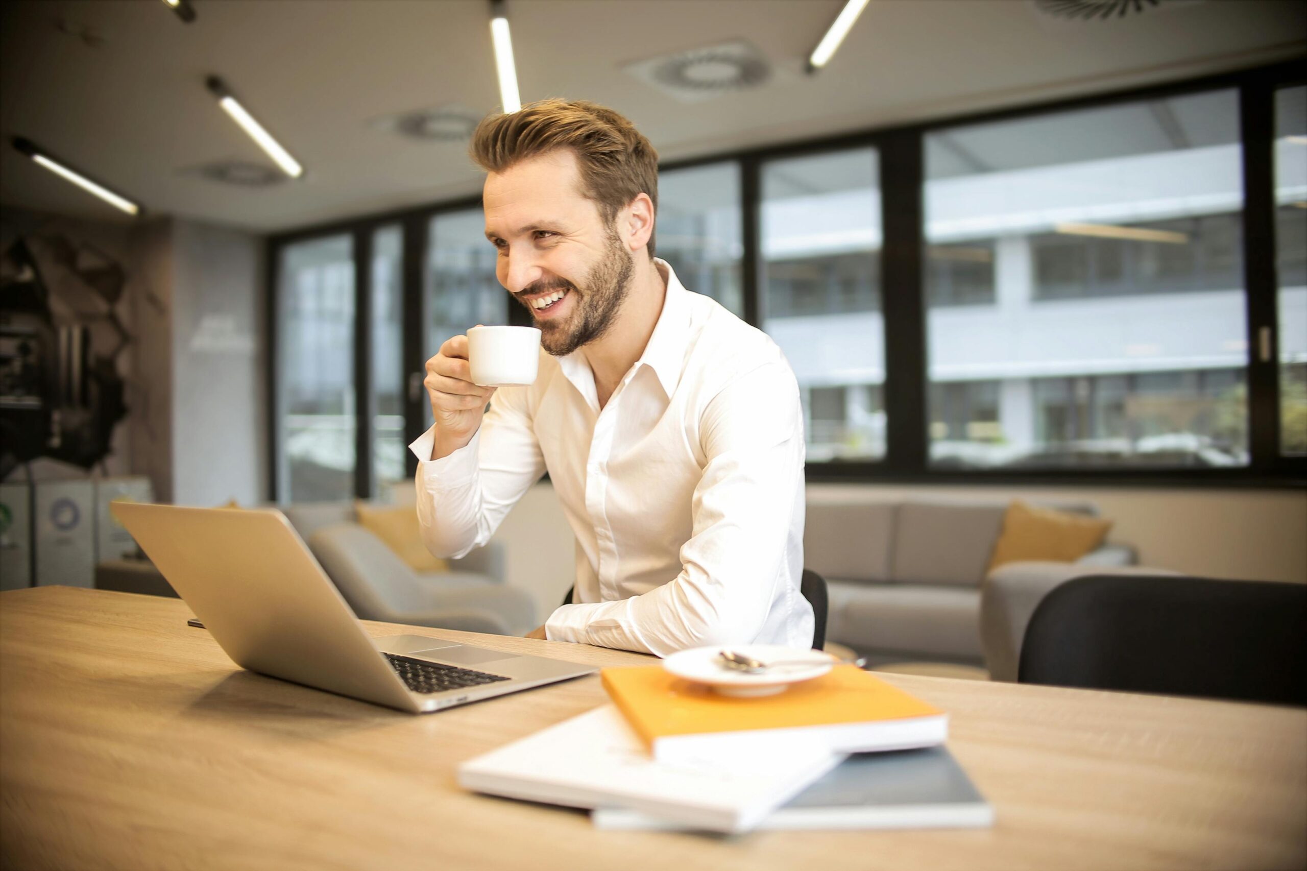 Hombre joven con barba, vestido con una camisa blanca, sonriendo mientras toma una taza de café frente a una computadora portátil en un ambiente de oficina moderno. En la mesa hay varios libros y una taza con plato.