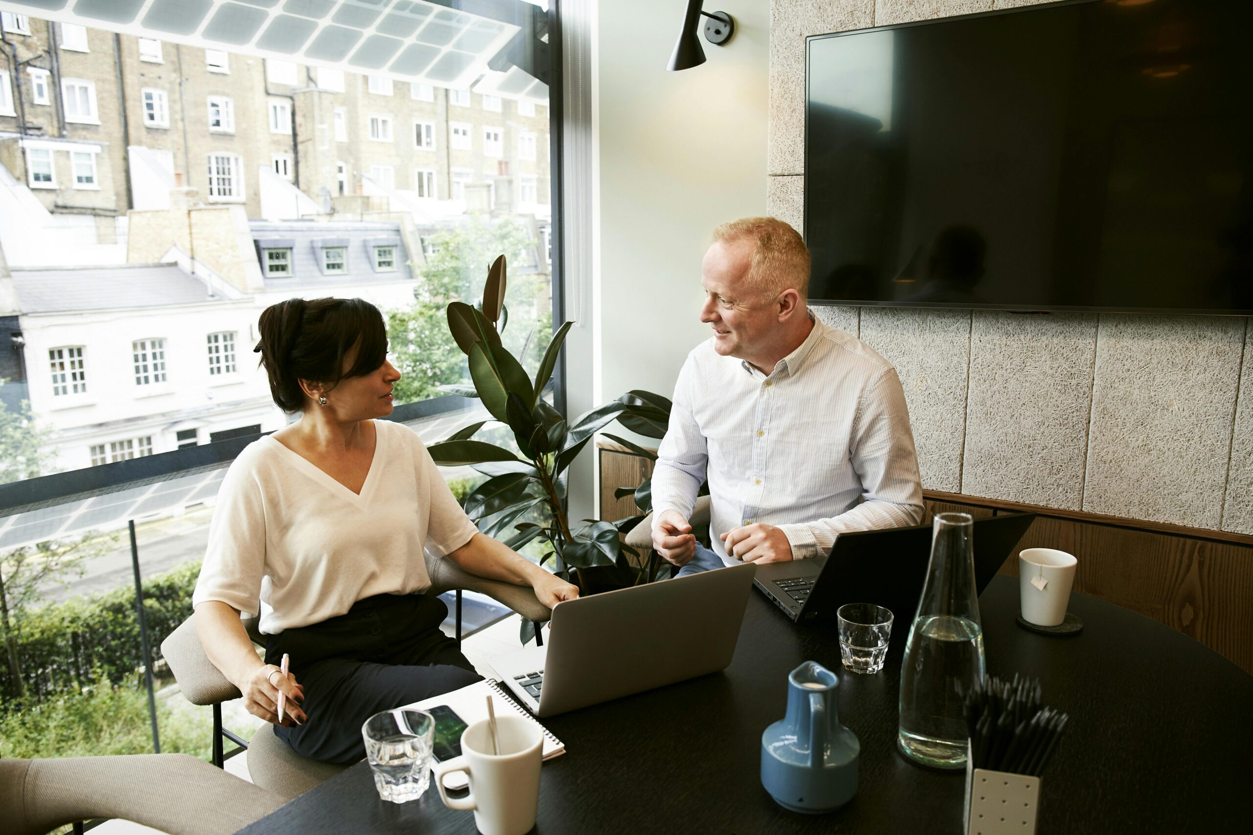 Two colleagues are having a discussion in a bright, modern office with large windows overlooking a cityscape. They are seated at a table with laptops, notebooks, and cups of coffee. The man is smiling and talking, while the woman listens attentively with a pen in her hand. There is a potted plant beside them, and a large screen is mounted on the wall behind them.