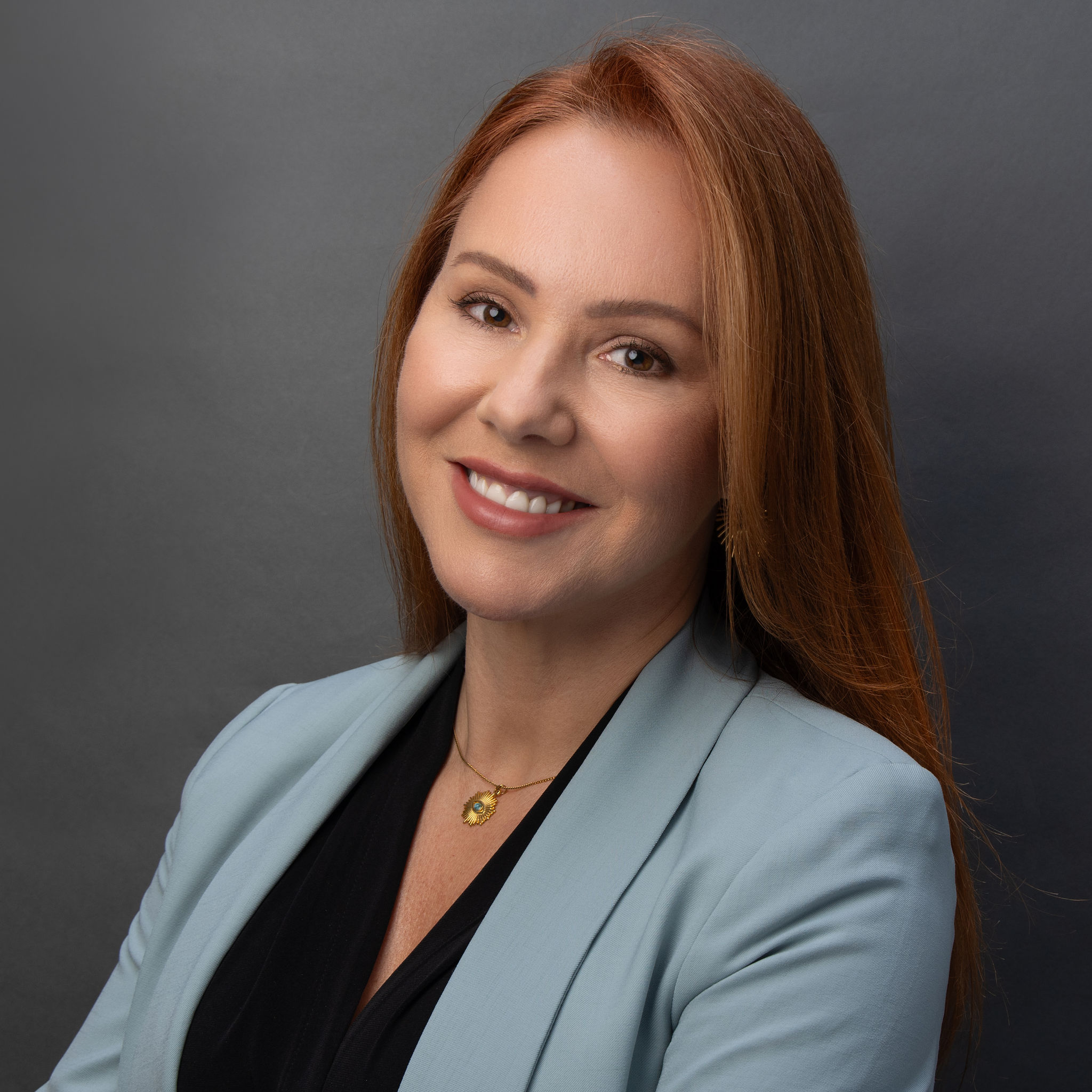 Professional headshot of a woman with long red hair, wearing a light blue blazer over a black top, smiling warmly against a neutral dark background.