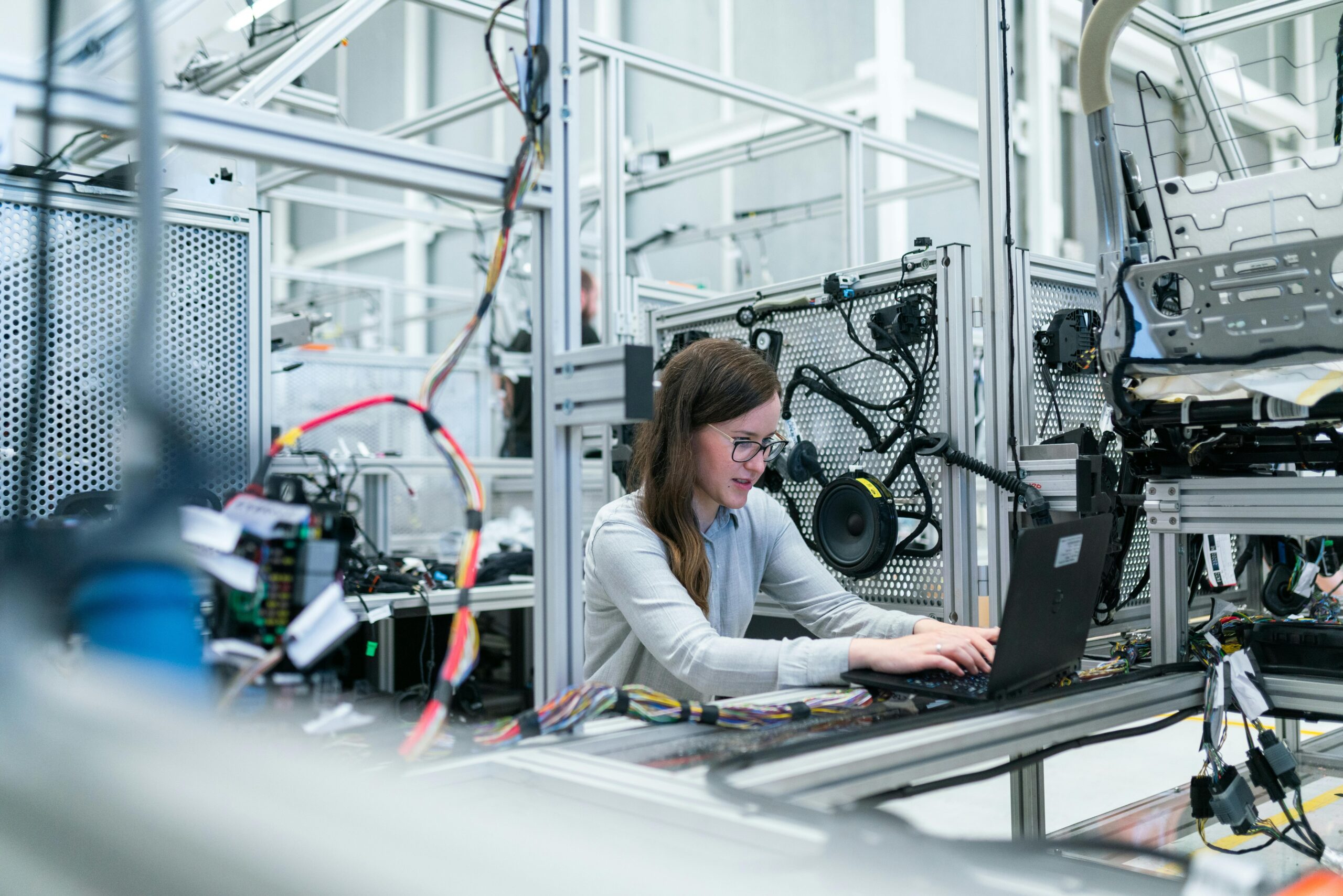 A female engineer working on a laptop surrounded by various electronic equipment and wiring in a high-tech laboratory or manufacturing facility