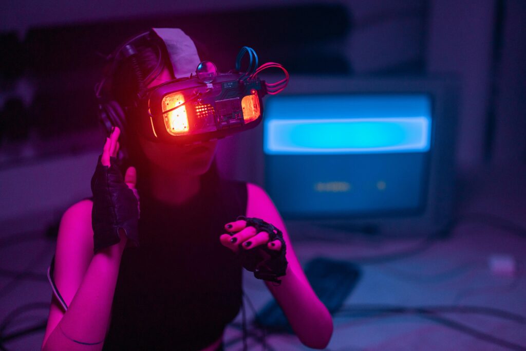 A person using a virtual reality headset with bright red lights, dark gloves, surrounded by a technological atmosphere with purple and blue lighting tones, and a computer screen in the background.