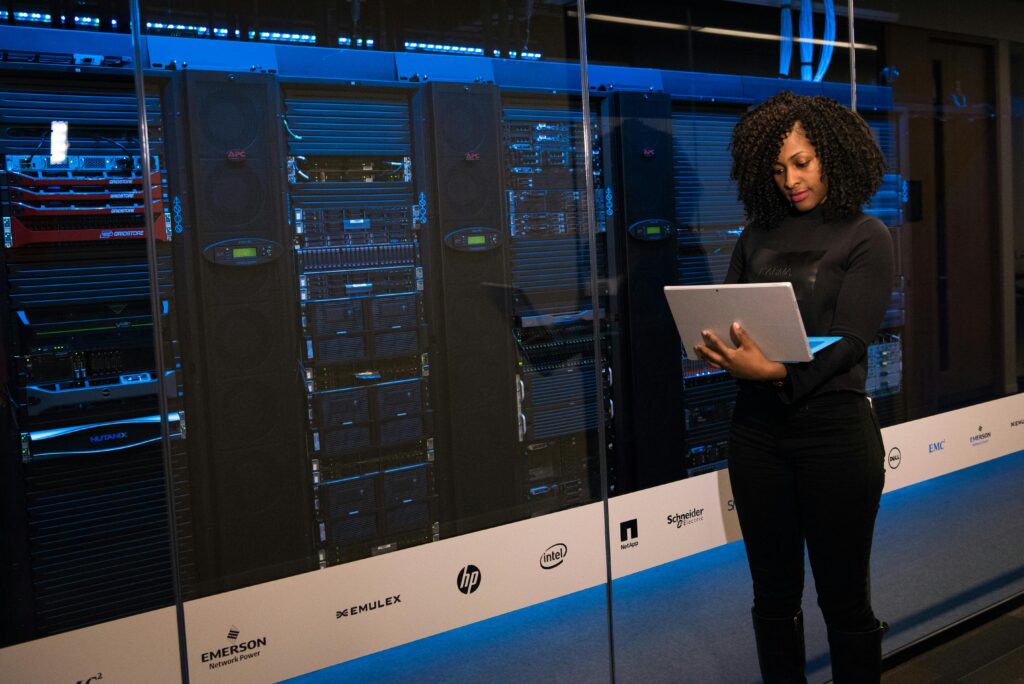 A professional woman with curly hair reviewing information on a laptop inside a server room, surrounded by advanced technological equipment, featuring renowned tech brands in a high-tech environment.