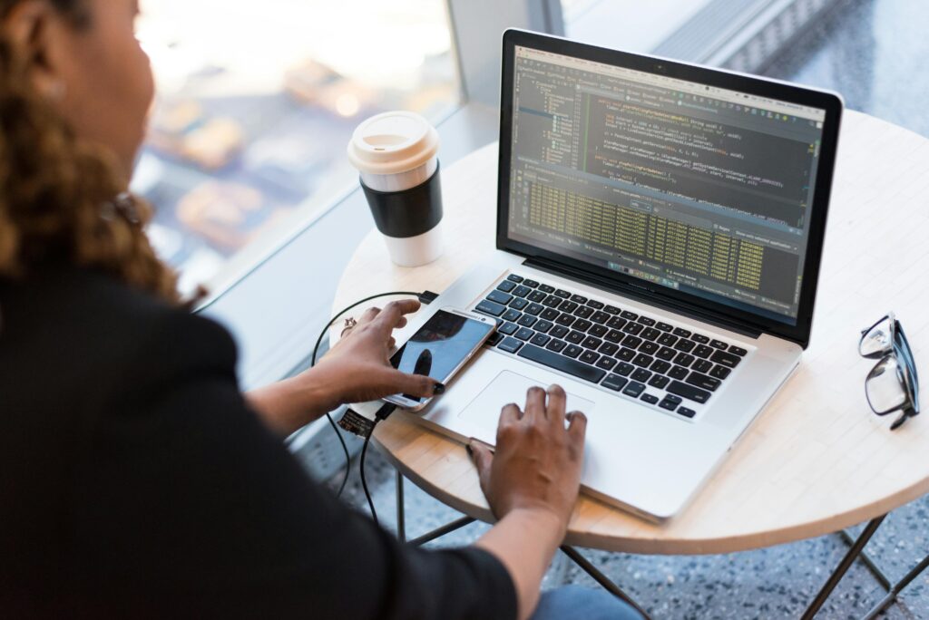 Mujer trabajando en una computadora portátil con código de programación en la pantalla, mientras sostiene un teléfono móvil en una mesa junto a una ventana con vista a la ciudad. Hay una taza de café y un par de gafas sobre la mesa.