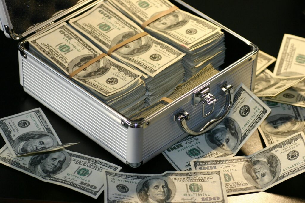 A metal briefcase filled with stacks of U.S. one hundred dollar bills, with additional bills scattered on the table.