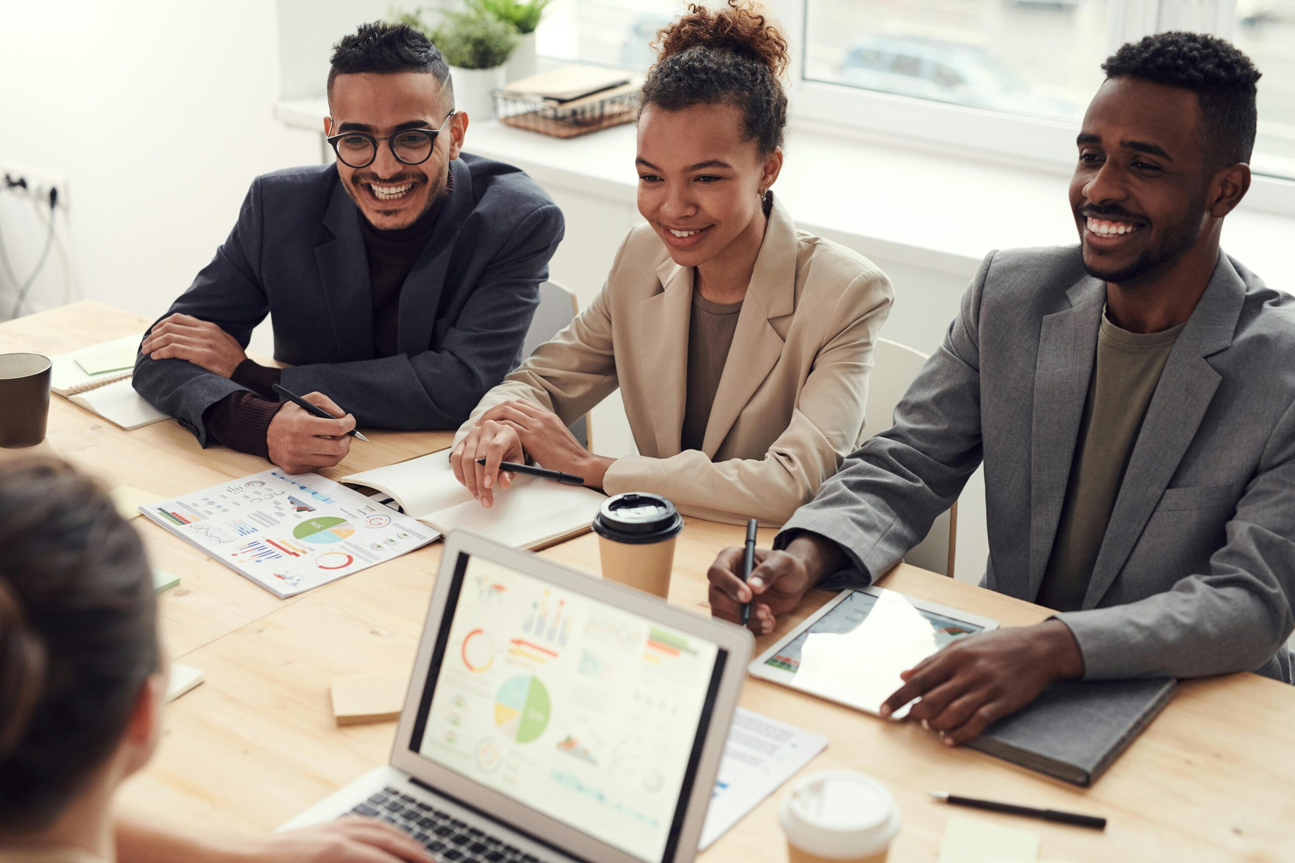 A group of smiling professionals sitting around a meeting table with laptops, charts, and coffee during a team discussion
