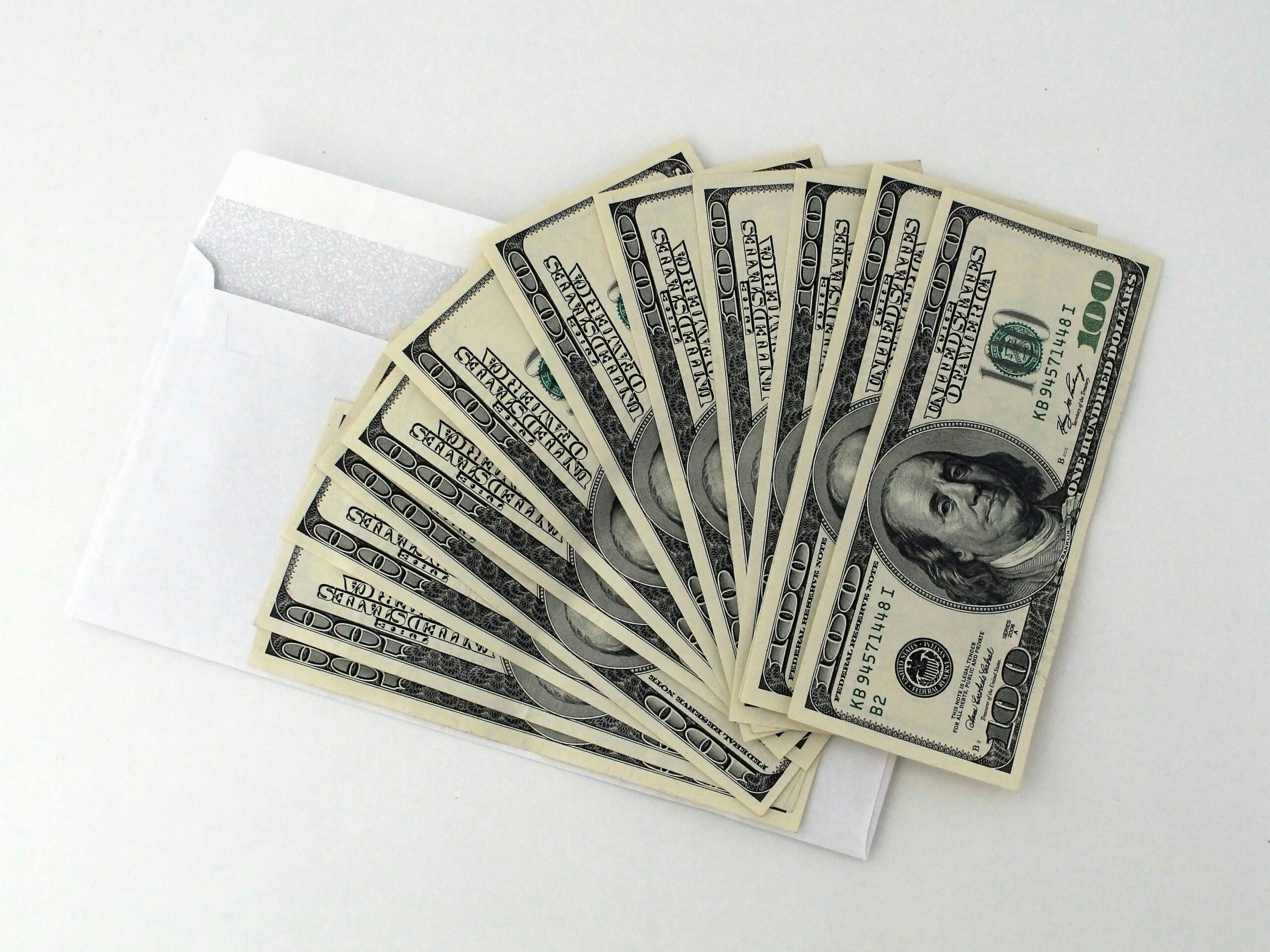 Un sobre blanco parcialmente abierto con varios billetes de 100 dólares estadounidenses extendidos en abanico sobre una superficie blanca