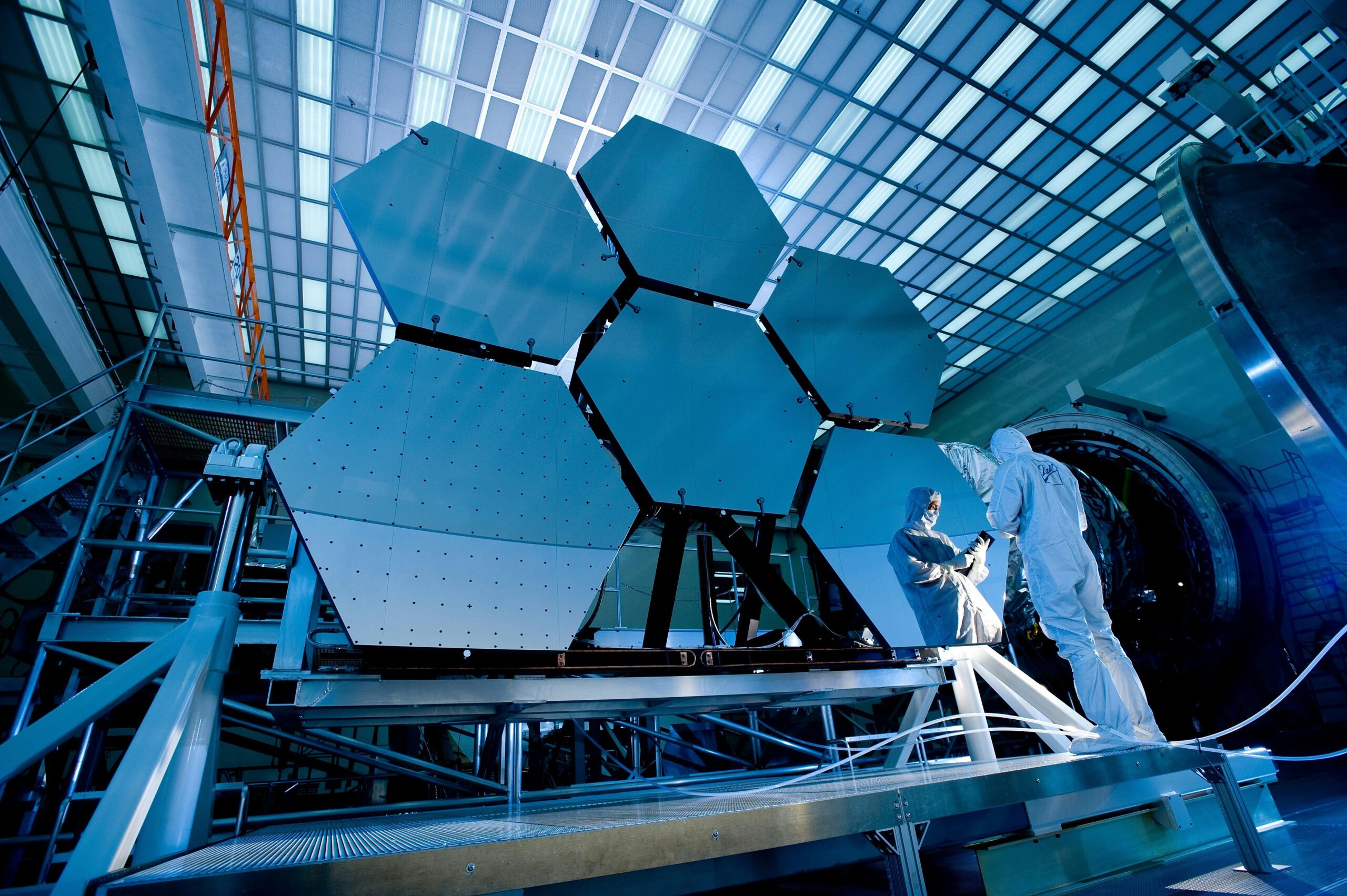 Two technicians in cleanroom suits inspecting a large hexagonal mirror array inside a high-tech laboratory or aerospace facility.