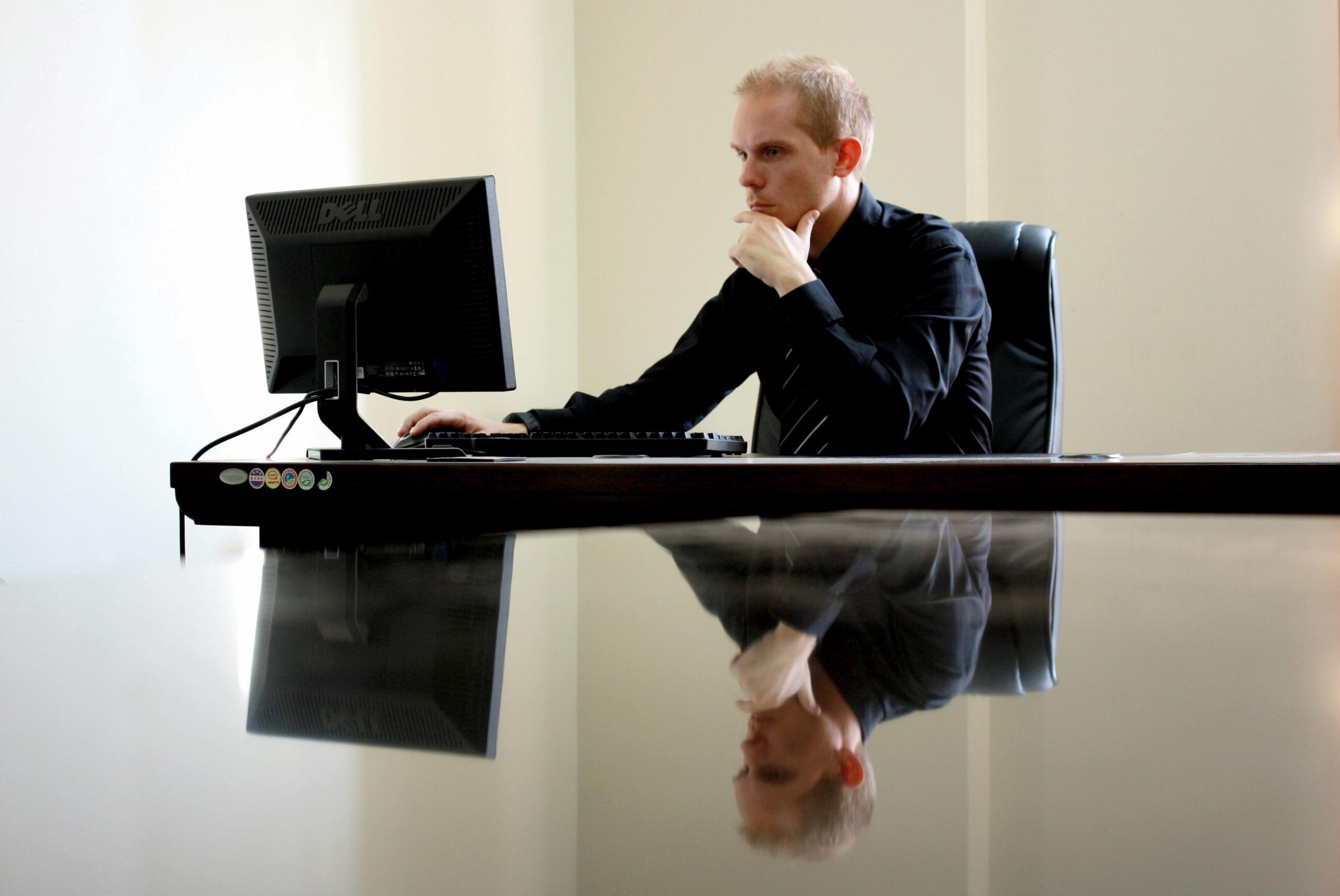 Business professional sitting at a desk in a modern office, working on a desktop computer with a thoughtful expression, reflected on a glossy table surface.